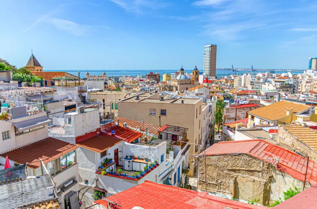Panoramic view from Barrio de Santa Cruz across Alicante's historic rooftops, harbour, and Mediterranean Sea.